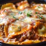 Close-up of a dish featuring ravioli in a tomato-based sauce, topped with melted cheese and garnished with fresh parsley. The ravioli, reminiscent of a simple slow cooker lasagna, are filled with a meat mixture, creating a warm and savory Italian meal in a black serving dish.