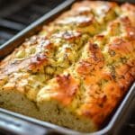 Close-up of freshly baked dill pickle bread in a rectangular pan. The bread has a golden-brown crust and is topped with herbs, capturing the tangy texture and warmth of homemade baking as it rests on the stovetop.