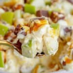 A close-up of a spoonful of creamy Snickers Salad showcases chunks of green apples and chocolate pieces, drizzled with caramel sauce. In the background, a bowl brims with this delectable Potluck Dessert.