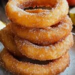 A stack of golden, sugar-coated apple cider donuts rests on a speckled gray plate, reminiscent of a delightful cinnamon apple dessert. In the background, sliced apples are partially visible on a wooden surface, evoking thoughts of cinnamon sugar apple rings.