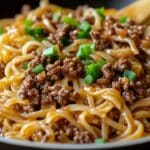 Close-up of a plate of Mongolian Ground Beef Noodles, topped with minced meat and chopped green onions. The noodles have a glossy appearance, suggesting a savory sauce coating. Wooden chopsticks rest on the side, ready for your first bite.