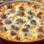 A close-up of a baked cheesy dip for chips featuring melted cheese and browned meatballs, garnished with chopped herbs. This easy beer cheese dip rests in a red round baking dish, while some crackers temptingly sit in the background.