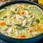 A bowl of traditional Greek soup featuring shredded chicken, rice, carrots, and lemon slices, garnished with dill. This Greek lemon chicken soup is served in a blue bowl with a slice of bread and surrounded by fresh lemon wedges.