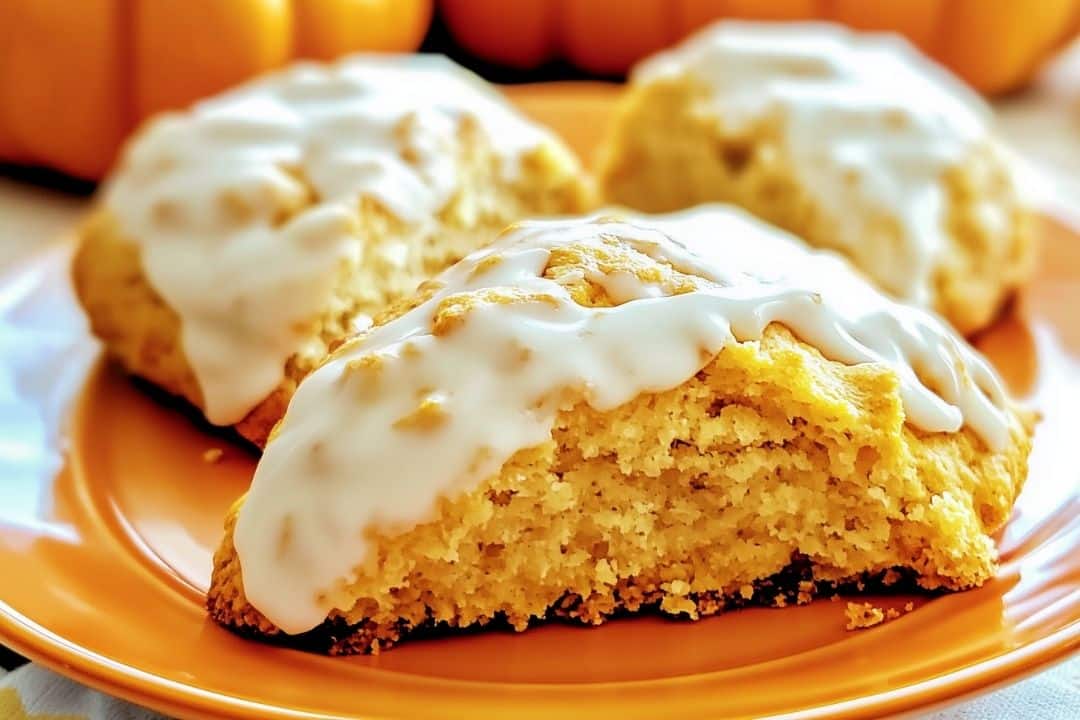 Three homemade pumpkin scones on an orange plate, boasting a golden-brown crumb and white icing atop. The background is softly blurred, beautifully highlighting the scones' texture and pumpkin-infused glaze.