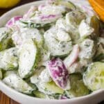 A bowl of dill cucumber salad boasts sliced cucumbers, red onions, and a creamy dill-infused dressing. A wooden spoon is visible in the top right corner, while a hint of lemon graces the wooden background.