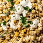 Close-up of a creamy Mexican Street Corn Pasta Salad with rotini, corn, black beans, and fresh cilantro. The dish is topped with dollops of sour cream and black pepper, served in a dark bowl.