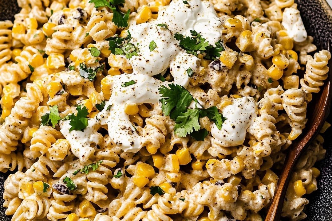 Close-up of a creamy Mexican Street Corn Pasta Salad with rotini, corn, black beans, and fresh cilantro. The dish is topped with dollops of sour cream and black pepper, served in a dark bowl.