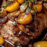 Close-up of a savory pot roast topped with caramelized onions and yellow cherry tomatoes, garnished with fresh thyme sprigs. The dish resembles a rich Apple cider braised pork recipe, highlighting the tender and juicy texture of the meat immersed in a luscious, brown sauce.