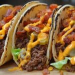 Three bacon cheeseburger tacos packed with seasoned ground beef, shredded lettuce, sliced pickles, and crispy bacon pieces, drizzled with melted cheese inside charred folded tortillas. A quick taco recipe sure to satisfy your cravings for a classic favorite with a twist.