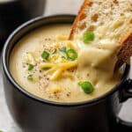 A bowl of creamy Roasted Garlic Cheddar Cauliflower Soup garnished with shredded cheese and chopped green onions. A slice of crusty bread is being dipped into the soup. The black bowl sits on a light-colored surface, complemented by an extra piece of bread in the background.