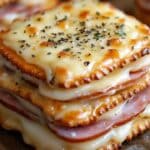 A close-up of savory crackers stacked with melted Swiss cheese and baked ham in between. The top is garnished with herbs and a golden, crispy layer of cheese. Perfectly described as Ham and Swiss Cracker Melts, they're a delightful homemade cracker melts recipe for any occasion.