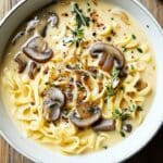 A bowl of creamy mushroom pasta garnished with fresh herbs and black pepper. The dish features wide, flat noodles reminiscent of an easy ramen noodles recipe, bathed in a rich, beige sauce, set on a wooden table.