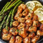 A skillet filled with Garlic Butter Chicken Bites, asparagus spears, and lemon slices, garnished with herbs. The chicken is golden brown with a shiny glaze, and the asparagus is bright green, contrasting beautifully with the zesty lemon slices.