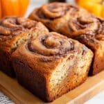 Four loaves of cinnamon swirl bread are displayed on a wooden board. Their golden-brown crusts reveal enticing swirls on top. Small pumpkins and cinnamon sticks blur in the background, enhancing the cozy, autumnal vibe. This small batch creation is perfect for pumpkin spice lovers.