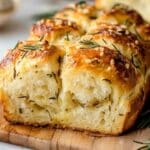 A loaf of golden-brown Rosemary Garlic Pull Apart Bread topped with rosemary sprigs and salt sits on a wooden board. This yeast bread is fluffy and textured, sliced to reveal its soft interior. Sprigs of rosemary are scattered around the board, enhancing its rustic charm.