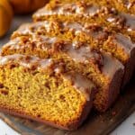 Sliced pumpkin bread with a glossy maple glaze sits on a wooden board. In the background, a small pumpkin and star anise add to the cozy autumn scene.