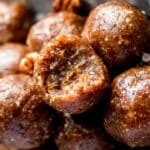 Close-up of several round, brown no-bake pecan pie energy bites with a glossy surface, topped with small pieces of nuts. Their sticky texture is inviting as they rest in a metal dish, making for an enticing no-bake dessert recipe.