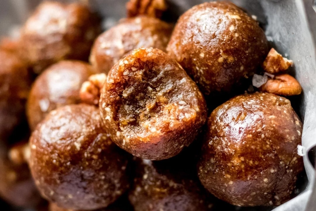 Close-up of several round, brown no-bake pecan pie energy bites with a glossy surface, topped with small pieces of nuts. Their sticky texture is inviting as they rest in a metal dish, making for an enticing no-bake dessert recipe.