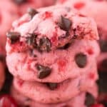 A stack of Maraschino Cherry Chocolate Chip Cookies, pink and studded with chocolate chips, sits enticingly with a bite missing from the top cookie. Bright red cherries surround the cookies, adding a vibrant touch to the scene.