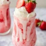 Two glasses filled with creamy strawberry milkshakes topped with whipped cream and fresh strawberries. The milkshakes, boasting a pink hue, feature streaks of fresh strawberry sauce. Additional strawberries are scattered on the table, evoking the perfect summer ice cream float mood.