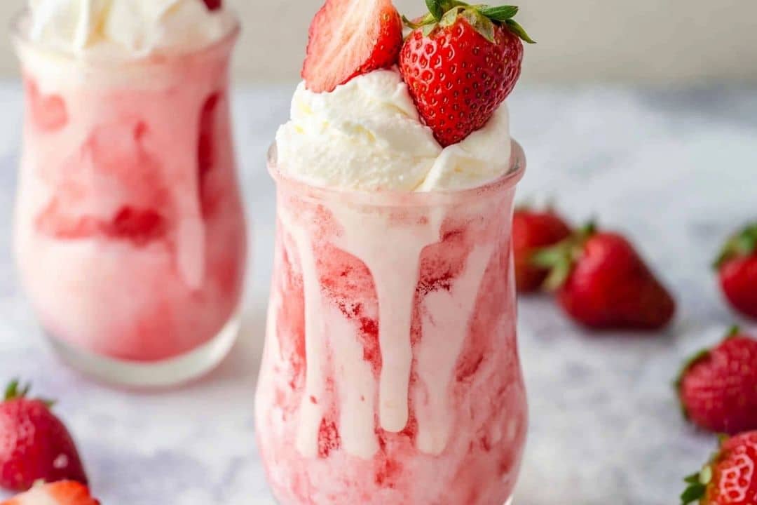 Two glasses filled with creamy strawberry milkshakes topped with whipped cream and fresh strawberries. The milkshakes, boasting a pink hue, feature streaks of fresh strawberry sauce. Additional strawberries are scattered on the table, evoking the perfect summer ice cream float mood.