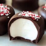 Close-up of chocolate-coated marshmallow treats topped with festive white and red sprinkles. One treat is halved, revealing the fluffy center reminiscent of Homemade Peppermint Patties.