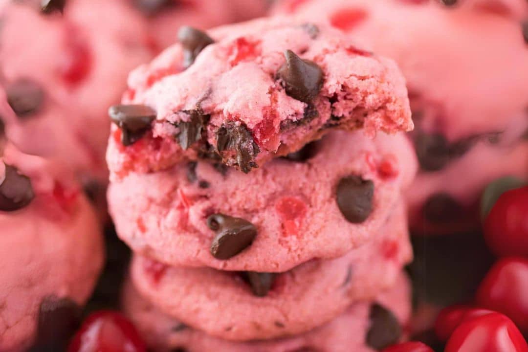 A stack of Maraschino Cherry Chocolate Chip Cookies, pink and studded with chocolate chips, sits enticingly with a bite missing from the top cookie. Bright red cherries surround the cookies, adding a vibrant touch to the scene.