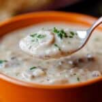 A close-up of a spoon lifting homemade creamy mushroom soup from an orange bowl. The soup, made with heavy cream, is garnished with chopped parsley, highlighting a rich and appetizing texture. A blurred background hints at bread and greenery.