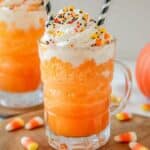 A frosty orange ice cream float with orange punch is topped with whipped cream and Halloween-themed sprinkles in a clear glass mug. Striped straws poke out, candy corn is scattered around, and a small pumpkin decorates the background—a perfect easy Halloween dessert.