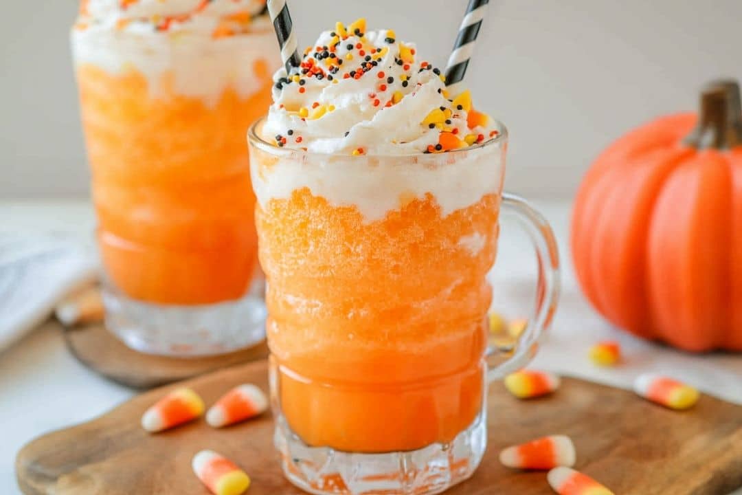 A frosty orange ice cream float with orange punch is topped with whipped cream and Halloween-themed sprinkles in a clear glass mug. Striped straws poke out, candy corn is scattered around, and a small pumpkin decorates the background—a perfect easy Halloween dessert.