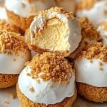 A plate of round, no-bake pumpkin cheesecake balls topped with white icing and crumbled biscuit topping. One treat has a bite taken out, revealing a creamy yellow filling inside.