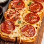 A close-up of a rectangular air fryer pizza toast with melted cheese and pepperoni slices on a wooden cutting board. The crust is thick and golden brown, sprinkled with herbs and black pepper. Bowls of dipping sauces are in the background.