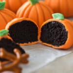 Miniature pumpkin-shaped cakes with orange icing and green leaves are displayed on parchment paper. These Halloween pumpkin treats reveal a dark chocolate sponge inside, resembling pumpkins. Brown candy sticks are artfully arranged in the foreground, adding rustic charm.