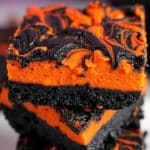 Two slices of Halloween swirl brownies, featuring an orange cheesecake layer with black swirl patterns, are artfully stacked on a plate. The dark brownie base perfectly complements the festive colors, making this a must-try Halloween swirl brownies recipe.