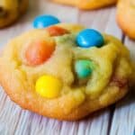 A colorful homemade M&M cookie with a soft, golden-brown texture is adorned with vibrant blue, yellow, and red candy-coated chocolates. The cookie is positioned on a light wooden surface, with additional candies scattered around.