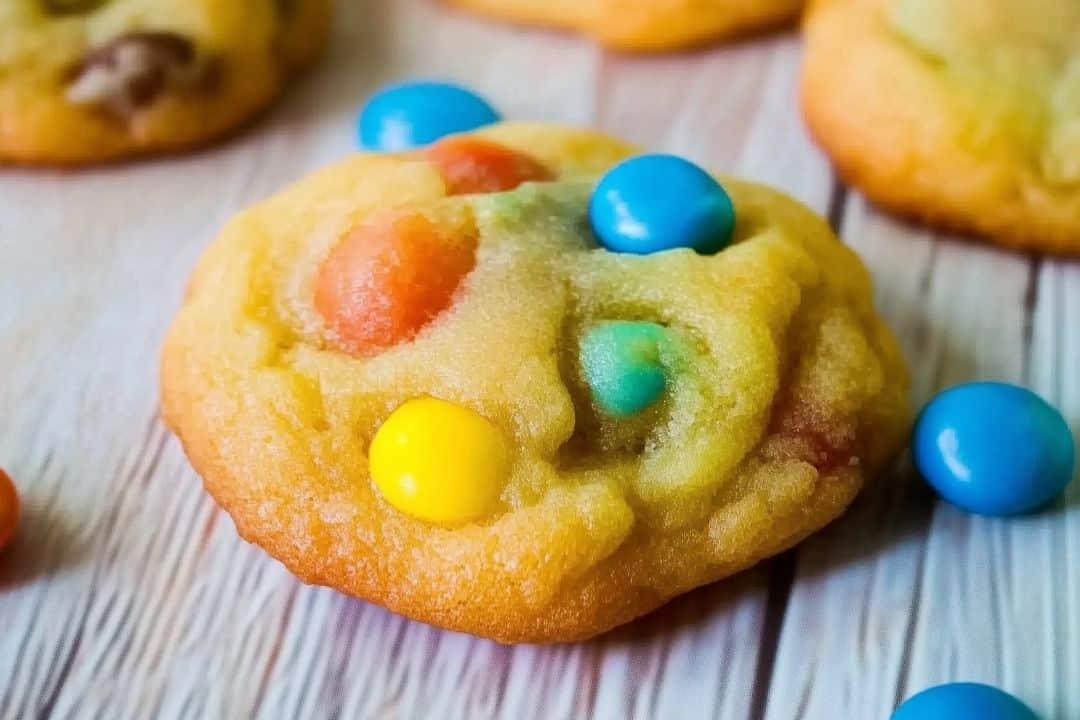 A colorful homemade M&M cookie with a soft, golden-brown texture is adorned with vibrant blue, yellow, and red candy-coated chocolates. The cookie is positioned on a light wooden surface, with additional candies scattered around.