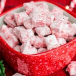 A red, speckled bowl filled with sugar-coated red and white candies sits on a red tablecloth. Sprigs of holly and a string of lights are in the background, enhancing the festive atmosphere. It almost feels like a scene from a holiday puppy chow recipe come to life.