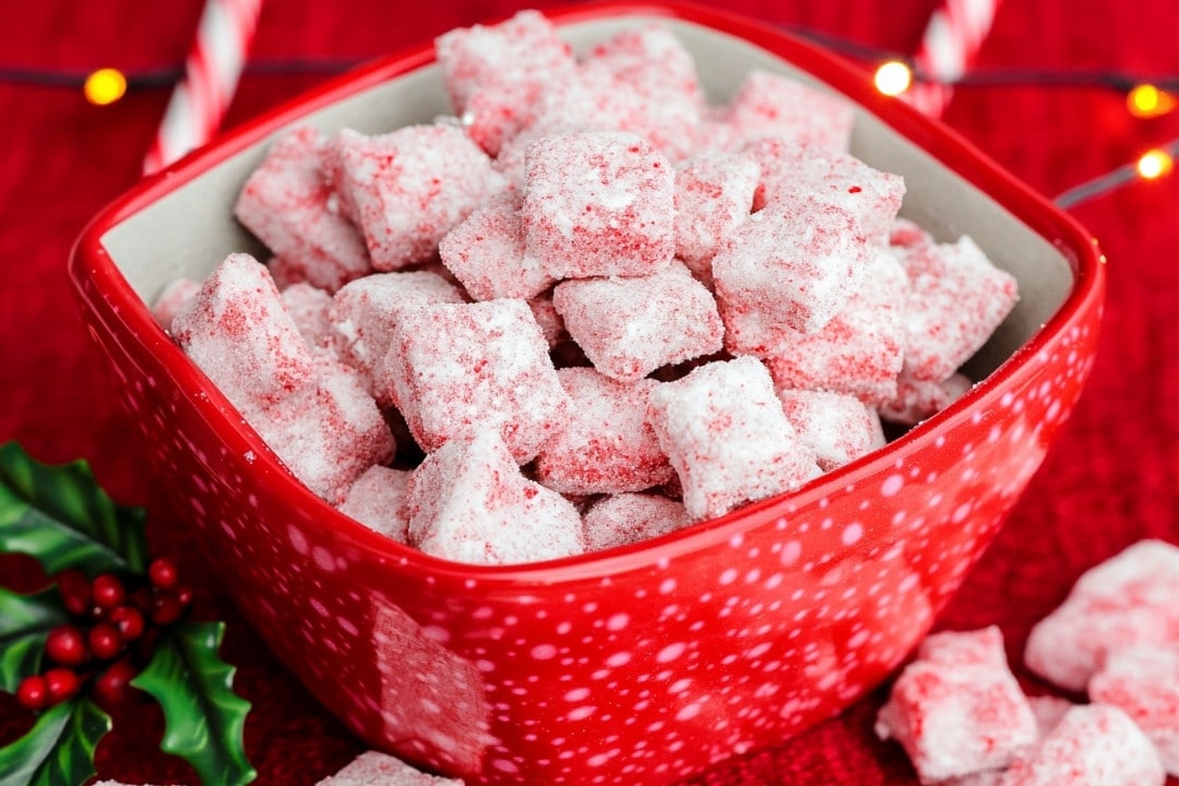 A red, speckled bowl filled with sugar-coated red and white candies sits on a red tablecloth. Sprigs of holly and a string of lights are in the background, enhancing the festive atmosphere. It almost feels like a scene from a holiday puppy chow recipe come to life.