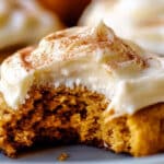 A close-up of an easy pumpkin cookie with cream cheese frosting, partially bitten to reveal the soft, moist interior. Topped with a sprinkle of cinnamon and surrounded by other frosted delights on a plate, this fall pumpkin cookie recipe is perfect for the season.