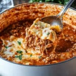 A close-up of a pot filled with creamy lasagna soup, rich and cheesy. A spoon lifts a portion, showcasing melted cheese, ground meat, and pasta. The dish is garnished with fresh herbs and sits on a rustic wooden table.