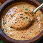 A wooden bowl filled with creamy, orange-colored queso dip garnished with red chili powder and chopped herbs. A hint of remoulade sauce for seafood flavor lingers as a spoon dips in, and tortilla chips are partially visible in the background.