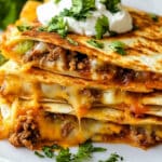 A stack of cheesy beef quesadillas, reminiscent of an easy cheeseburger quesadilla recipe, garnished with fresh cilantro and topped with a dollop of sour cream. Served with creamy guacamole on the side.