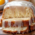 A moist spiced eggnog bread topped with a glossy glaze is sliced and displayed on a wooden board. In the background are cinnamon sticks and a bowl of fruit, adding cozy and fresh elements to this festive presentation.