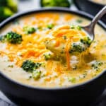 A black bowl filled with homemade broccoli cheese soup, garnished with shredded cheddar and broccoli florets. A spoon, holding a portion of the creamy soup, rests above the bowl. More broccoli and another bowl are in the blurred background.