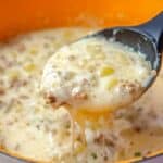 A ladle scoops creamy, cheesy hamburger potato soup from an orange pot. This 30-minute delight boasts ground beef, tender potatoes, corn, and visible melted cheese for a hearty, thick texture.