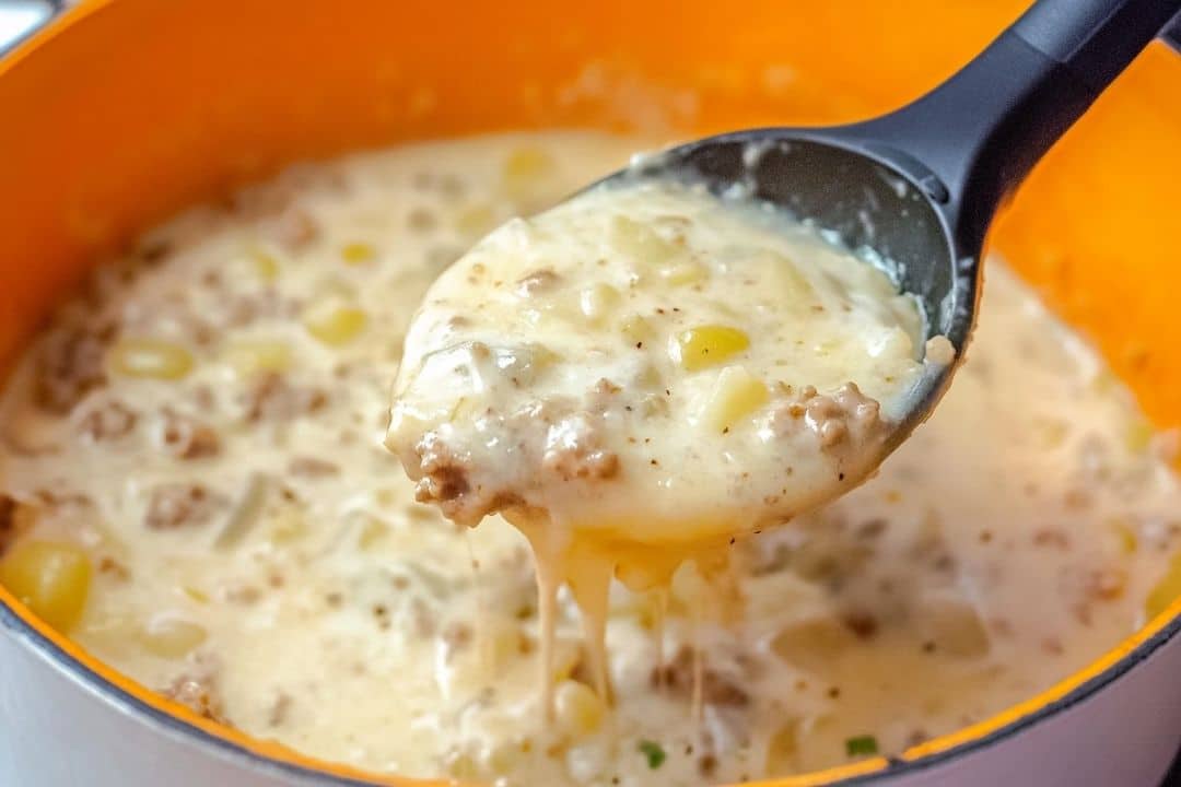 A ladle scoops creamy, cheesy hamburger potato soup from an orange pot. This 30-minute delight boasts ground beef, tender potatoes, corn, and visible melted cheese for a hearty, thick texture.