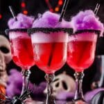 Three Halloween-themed cocktails, reminiscent of a Vampire Blood Halloween Drink, showcase vibrant red liquid and fluffy purple cotton candy on top. Served in glasses with black sugar rims, they're surrounded by skulls, a pumpkin, and a spider against glowing orange lights—perfect for your spooky Halloween party.