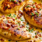 Close-up of a skillet with three browned chicken breasts in a creamy beer cheese sauce, garnished with crispy bacon bits and chopped chives. This delectable beer cheese chicken looks savory and well-seasoned, with a rich, golden sauce.