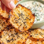 A hand holds a crispy cottage cheese chip covered with sesame seeds above a bowl of creamy dip garnished with herbs. More baked cottage cheese chips surround the bowl, creating a savory snack arrangement.