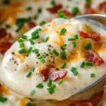 Close-up of a spoonful of creamy potato bacon soup topped with crispy bacon bits, shredded cheese, and chopped chives. The soup is light in color and appears rich and hearty, with visible seasoning and garnishes.